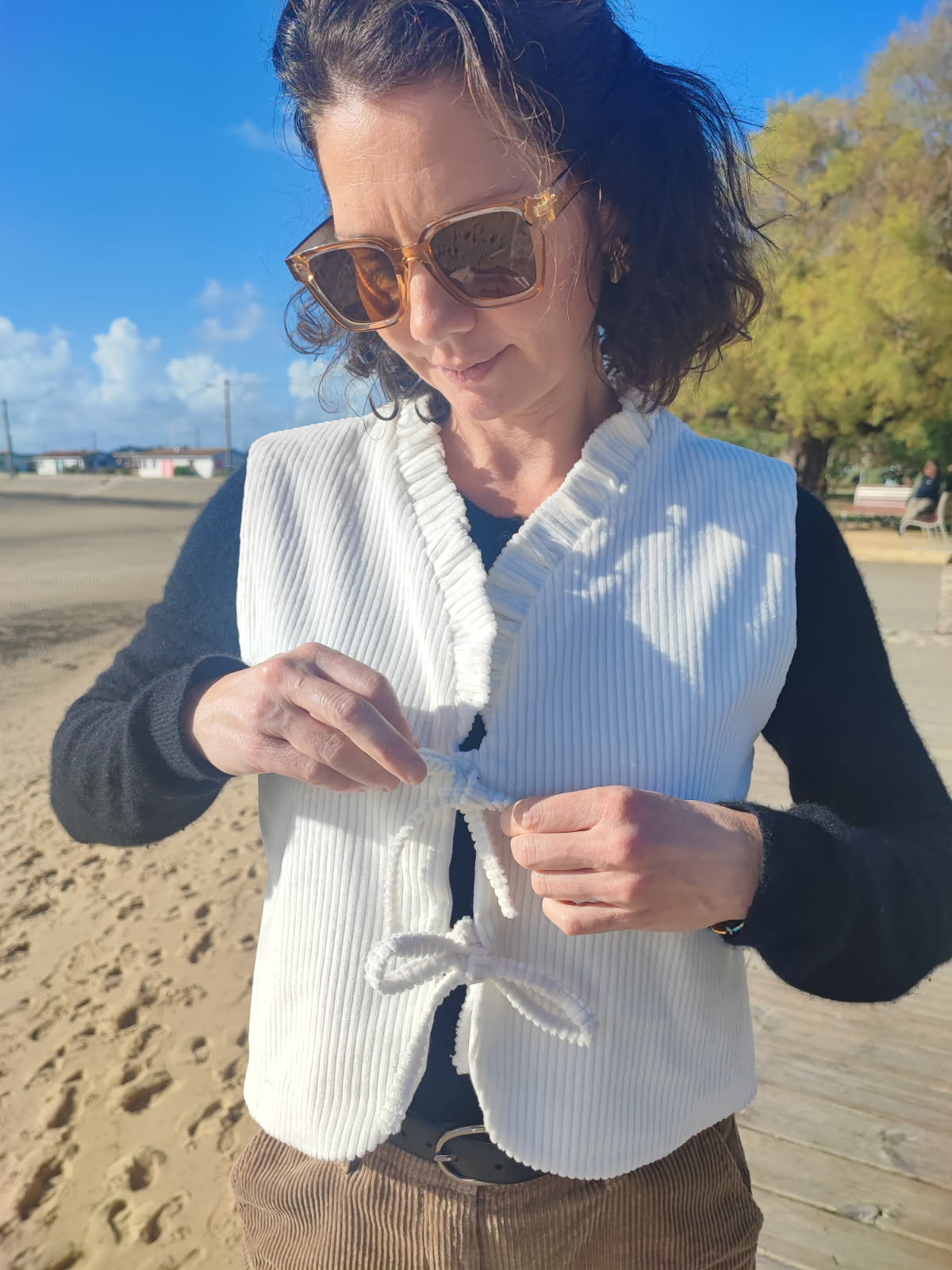 Gilet sans manches Cosmos en velours côtelé blanc cassé vu de face.
Le modèle est en train de nouer le gilet à l'aide de liens.